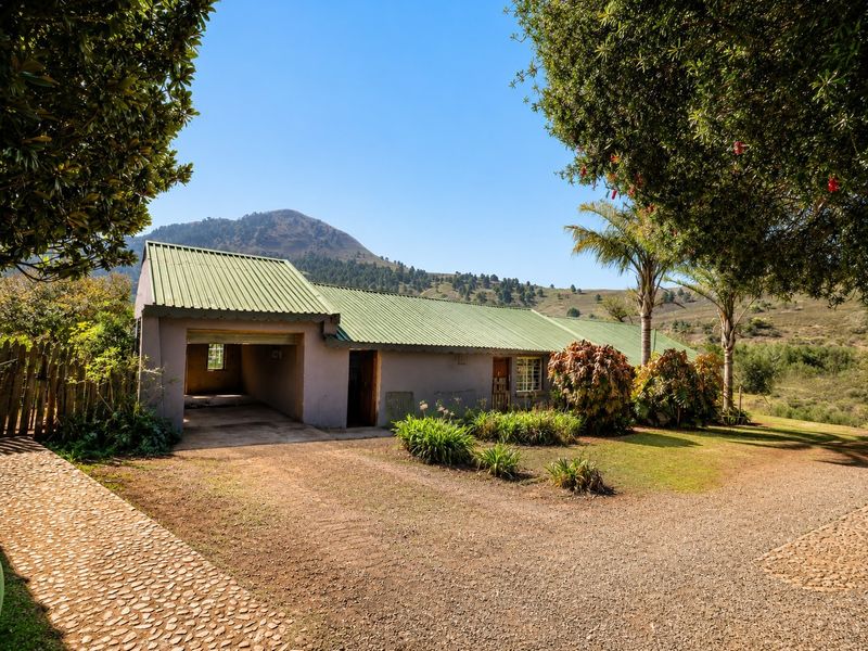Front view of a farm-style residence featuring a corrugated metal roof, attached garage, and gravel driveway set against a scenic mountain landscape.
