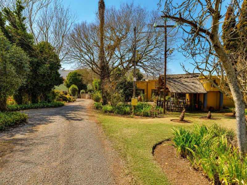 A scenic view of the property entrance featuring a gravel driveway, mature trees, landscaped garden areas, and a building with a traditional thatched roof.