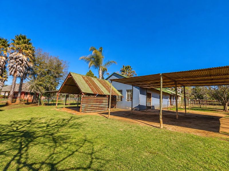Rear view of the farm property featuring a large open-sided shelter, a carport structure, and expansive lawn area under a clear blue sky.