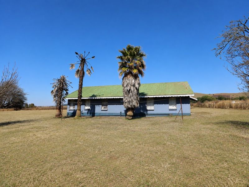 A rear view of a single-story farm cottage featuring a corrugated metal roof and security bars on the windows, set on a large open plot.