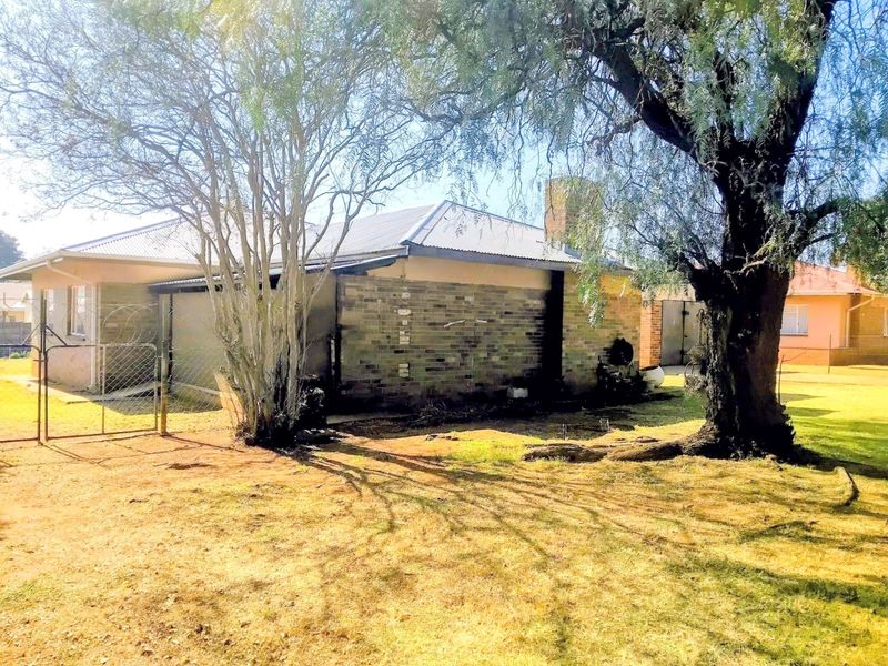 Rear view of the property featuring a brick facade, metal roof, chimney, and established trees in the garden area.