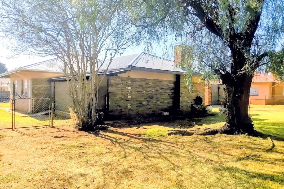 Rear view of the property featuring a brick facade, metal roof, chimney, and established trees in the garden area.