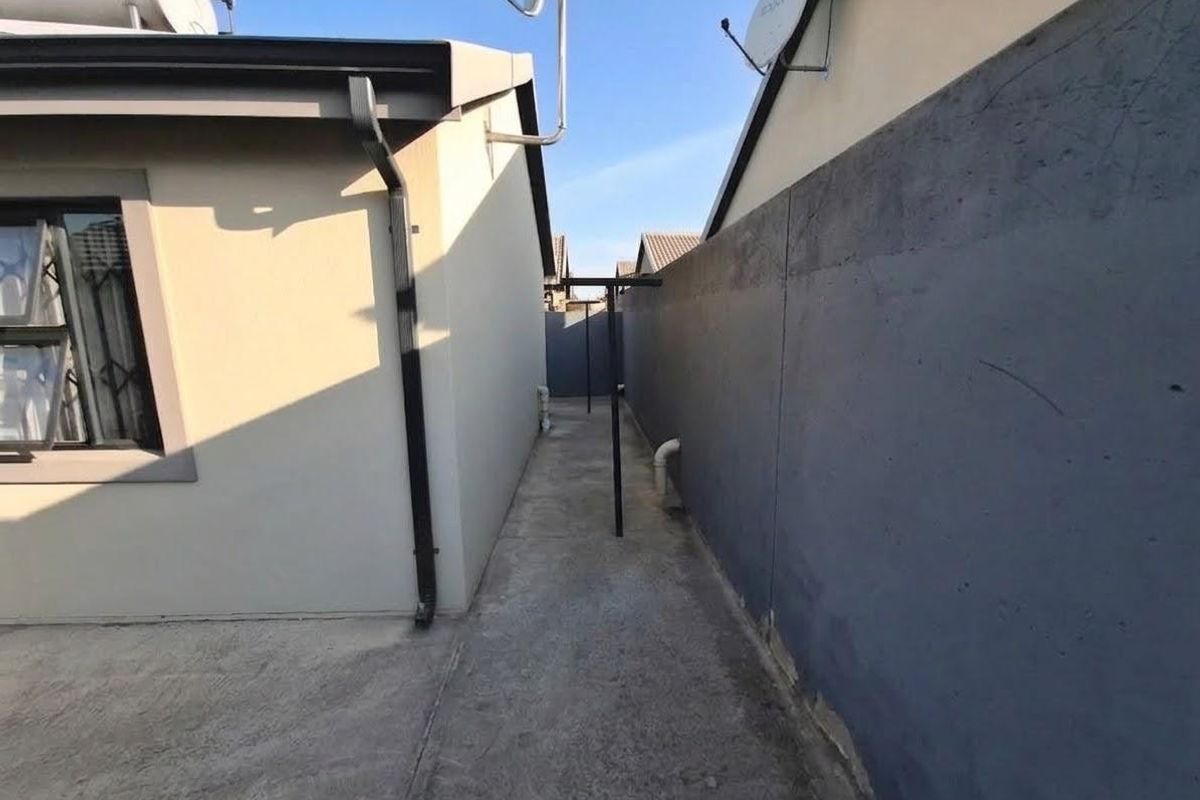 A narrow side passage featuring concrete paving, a boundary wall, and exterior utility installations including a solar geyser and satellite dish.
