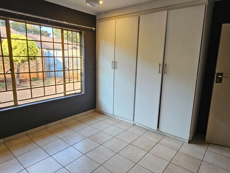 A bright, empty bedroom featuring tiled flooring, built-in cupboards, and a large window with security bars overlooking the exterior.