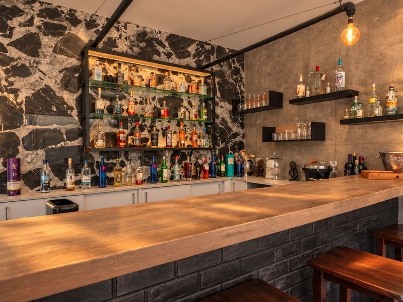 A stylish, well-appointed home bar featuring a solid timber counter, marble feature wall, and custom floating shelving with integrated lighting.