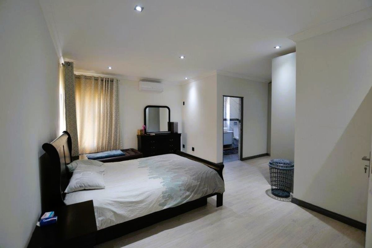 Spacious bedroom featuring light-toned laminate flooring, recessed downlighting, and an integrated air conditioning unit, with an adjoining en-suite bathroom.