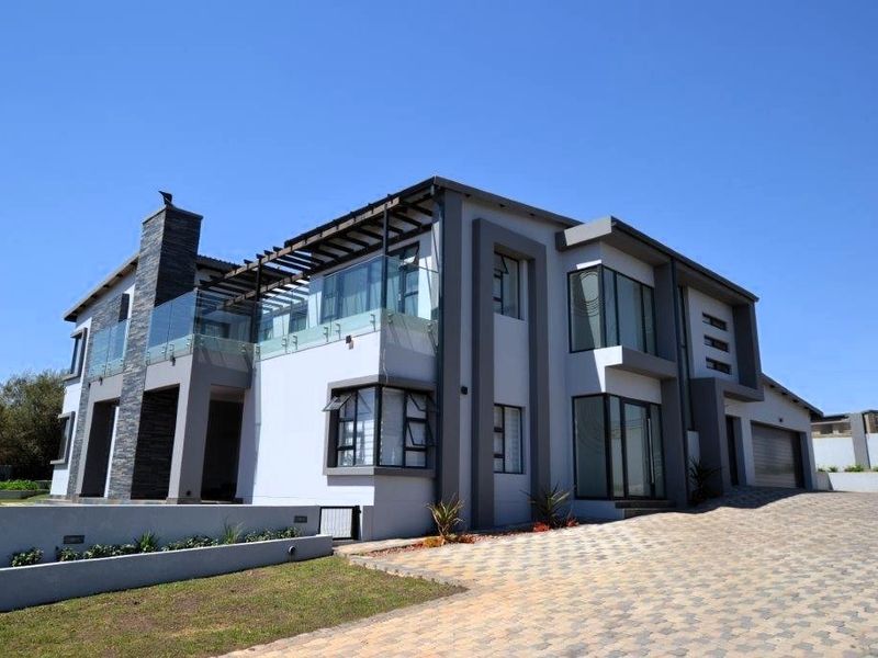 Modern double-storey residence featuring a stone-clad chimney, glass balcony balustrades, timber pergola, and a spacious paved driveway leading to a double garage.