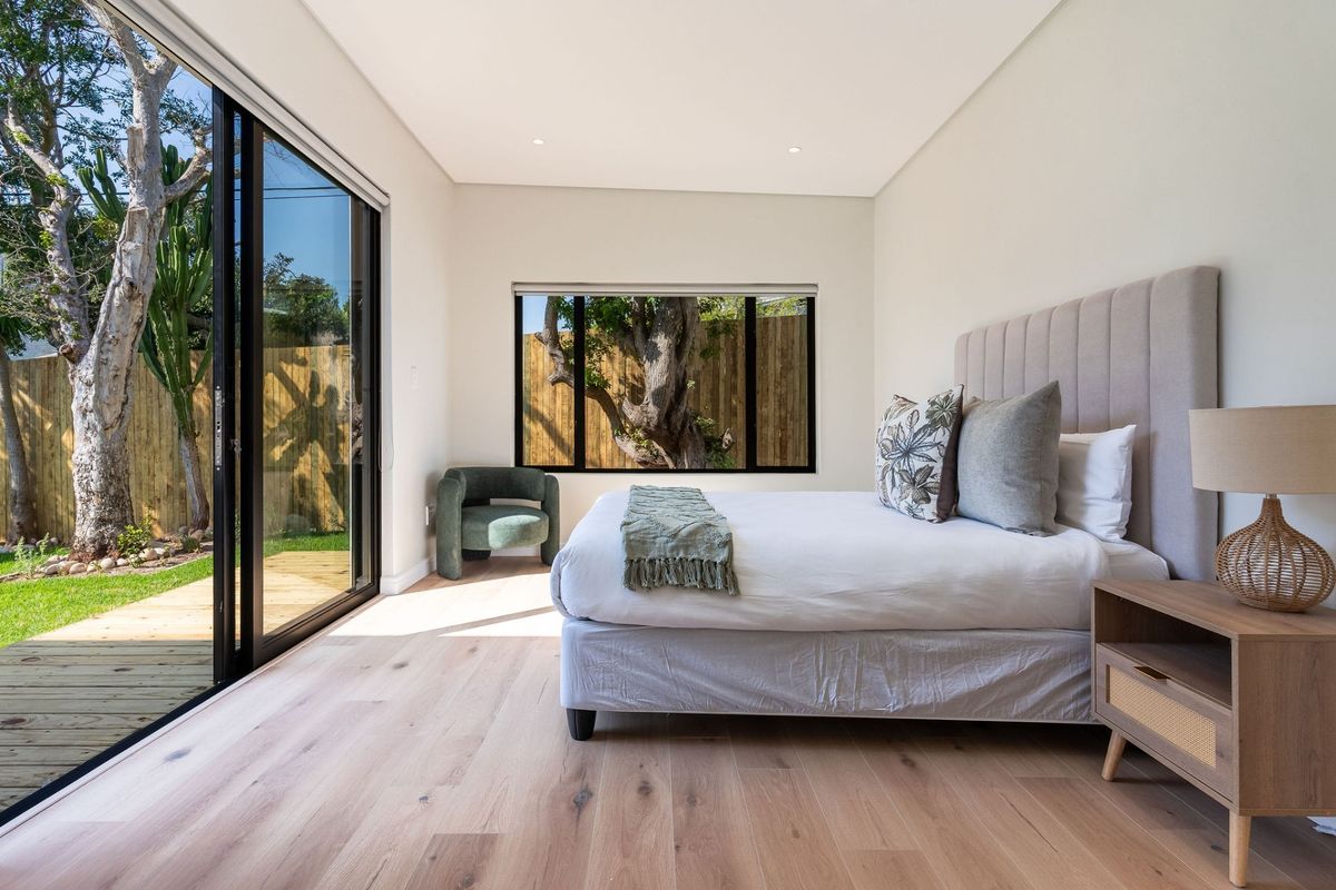 A bright, modern bedroom featuring light timber flooring, recessed lighting, and large sliding glass doors that open onto a private garden area.