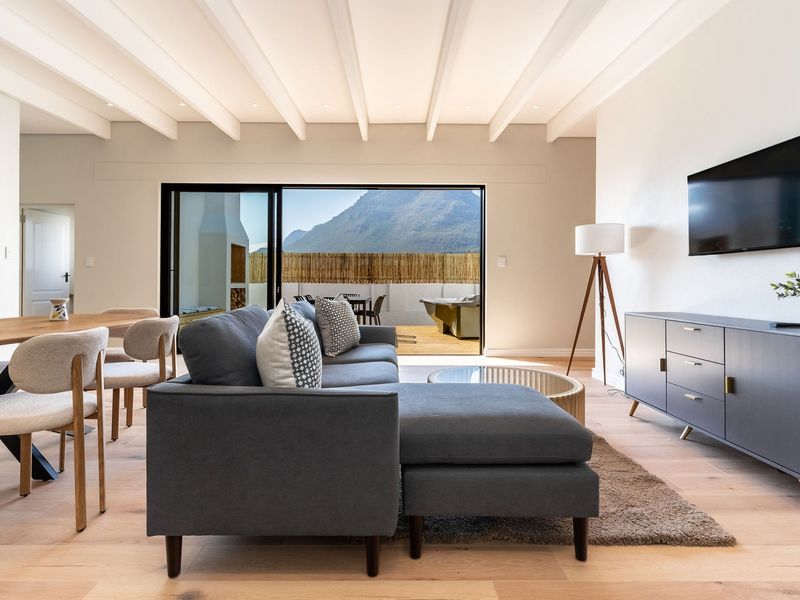 Bright open-plan living and dining area featuring exposed beam ceilings, light hardwood flooring, and large sliding doors opening to an outdoor patio with mountain views.