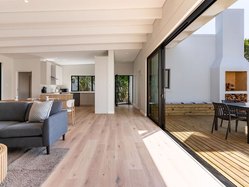 Bright open-plan living area featuring light hardwood floors, exposed beams, and seamless flow to a timber deck with a built-in outdoor fireplace.