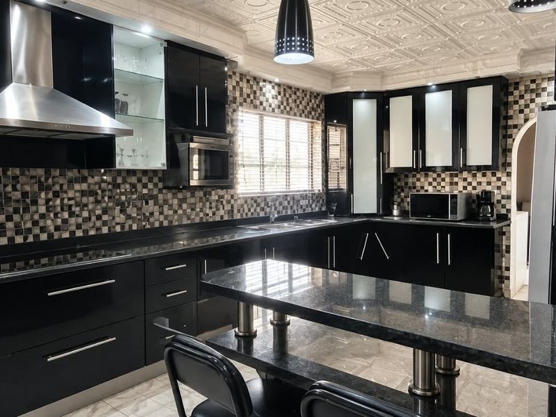 Modern kitchen featuring high-gloss black cabinetry, granite countertops, a breakfast bar, and decorative ceiling tiles with integrated stainless steel appliances.