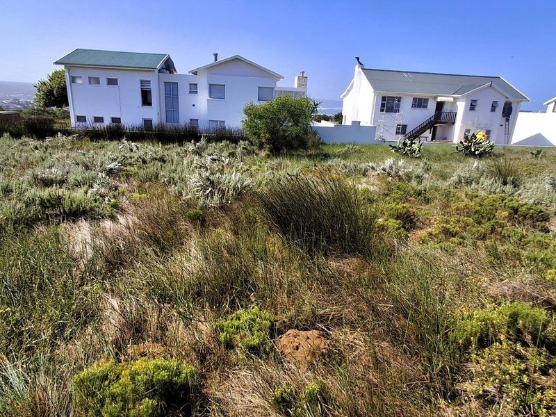 A view of a vacant residential plot featuring natural fynbos vegetation with neighboring residential properties visible in the background.