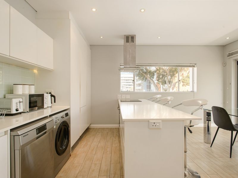 Modern open-plan kitchen featuring sleek white cabinetry, stone countertops, integrated appliances, and a central island with breakfast bar seating.