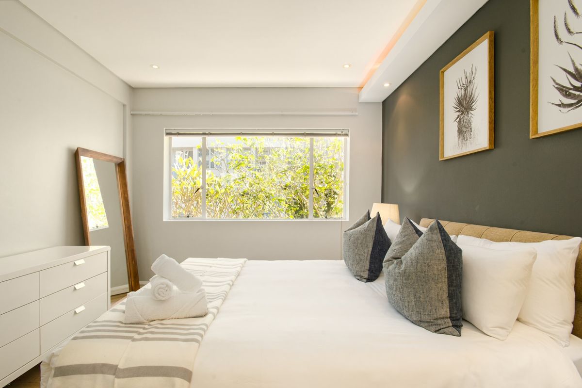 A well-lit, modern bedroom featuring a neutral color palette, recessed ceiling lighting, and a large window overlooking greenery.