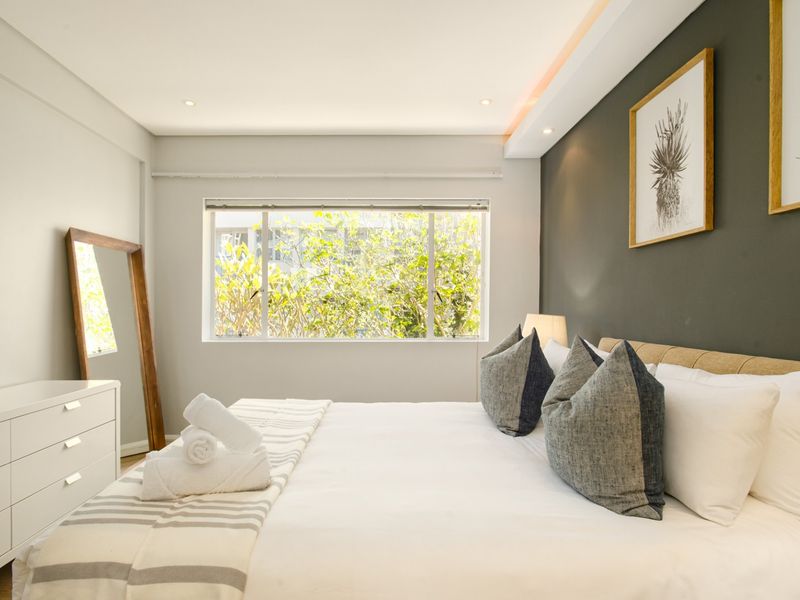 A well-lit, modern bedroom featuring a neutral color palette, recessed ceiling lighting, and a large window overlooking greenery.