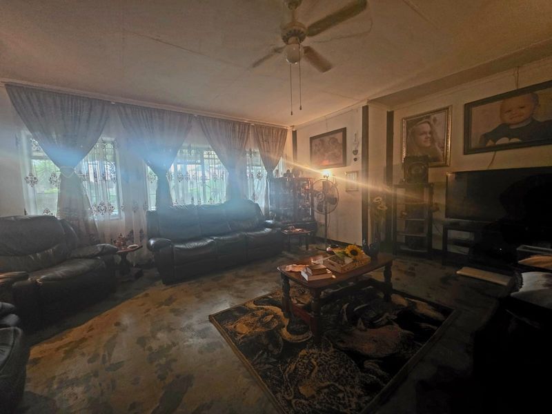 A living room featuring a ceiling fan and large windows. The space shows signs of wear on the flooring and ceiling finishes.