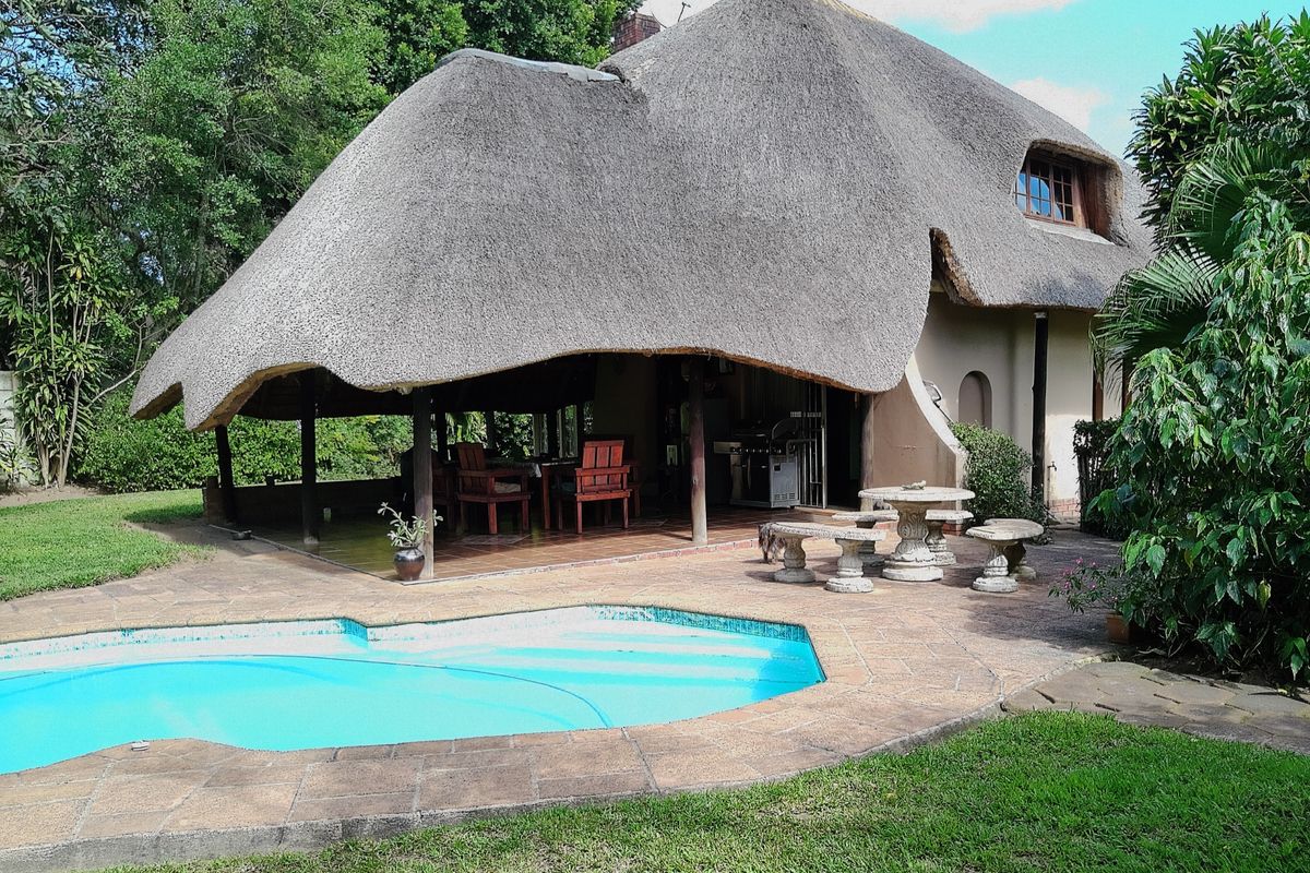 Rear view of a unique thatched-roof home featuring a covered patio entertainment area and a swimming pool set within a landscaped garden.