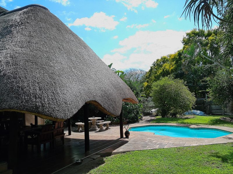 Outdoor entertainment area featuring a thatched lapa, paved patio, and a swimming pool set within a landscaped garden.