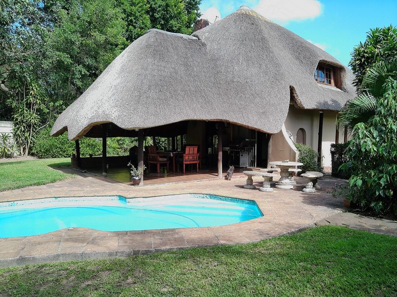 Rear view of a thatched-roof house featuring a covered patio area, paved outdoor space, and a swimming pool set within a garden.