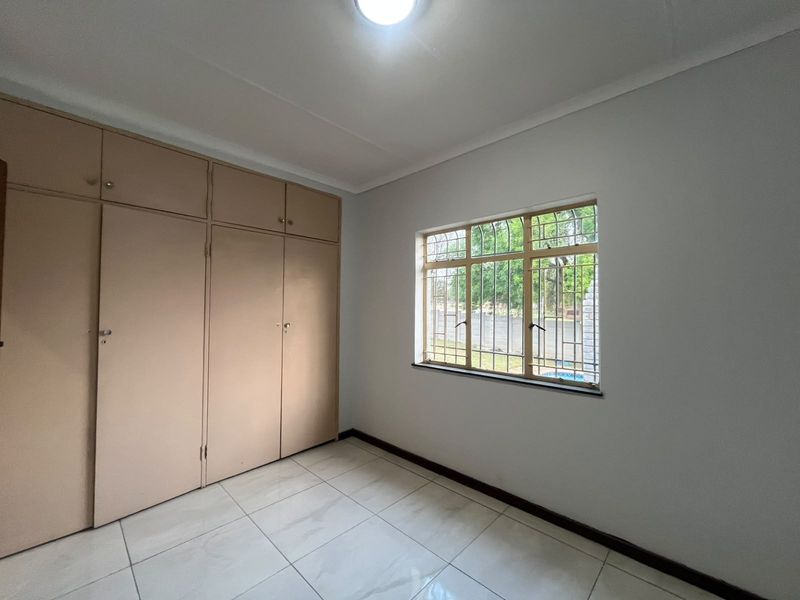 A clean, empty bedroom featuring neutral tiled flooring, built-in cupboards for storage, and a window fitted with security bars.