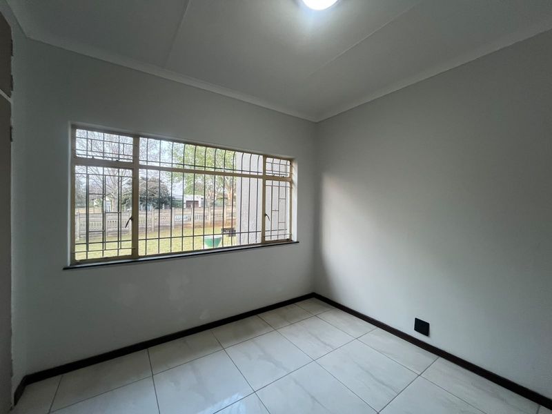 A bright, empty bedroom featuring neutral tiled flooring, clean walls, and a large window fitted with security bars.