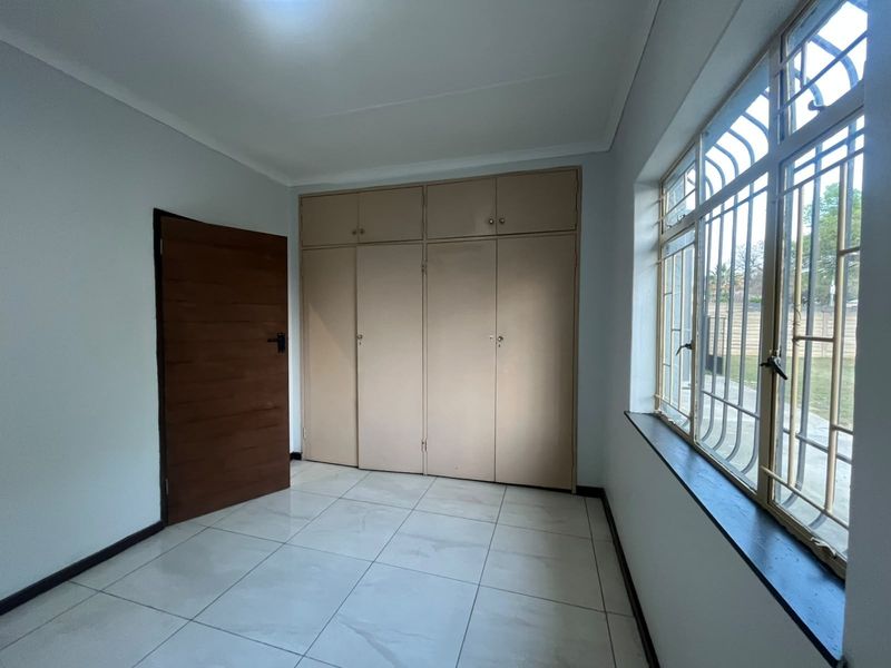 A bright, empty bedroom featuring tiled flooring, built-in cupboards, and large windows fitted with security bars.