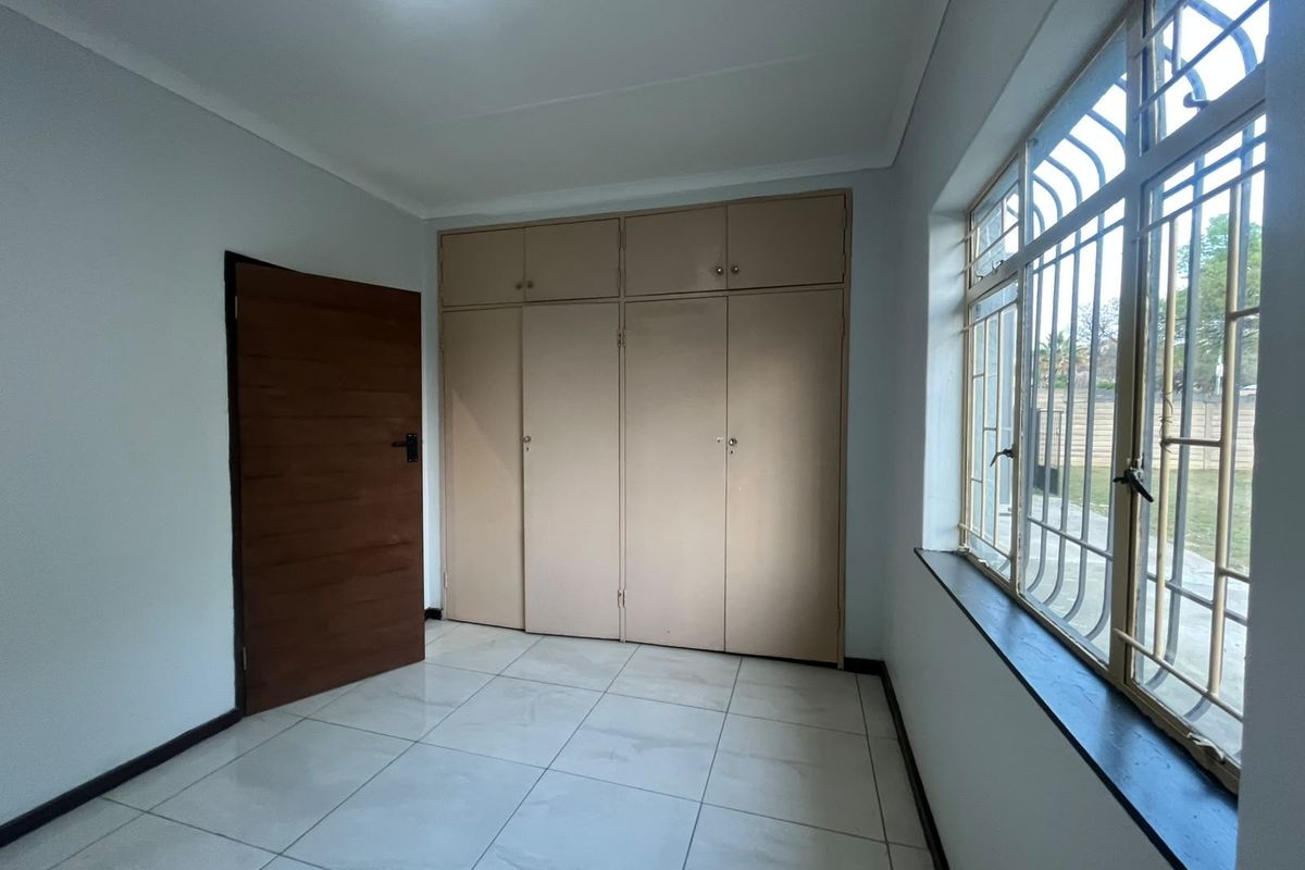 A bright, empty bedroom featuring tiled flooring, built-in cupboards, and large windows fitted with security bars.