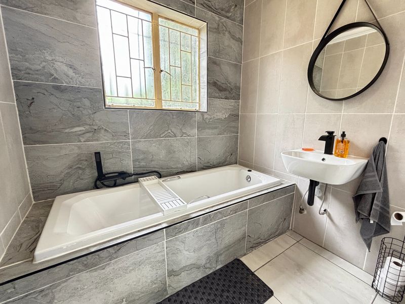 Modern bathroom featuring a built-in bathtub with grey stone-look tiling, a wall-mounted basin with black fixtures, and large format floor tiles.