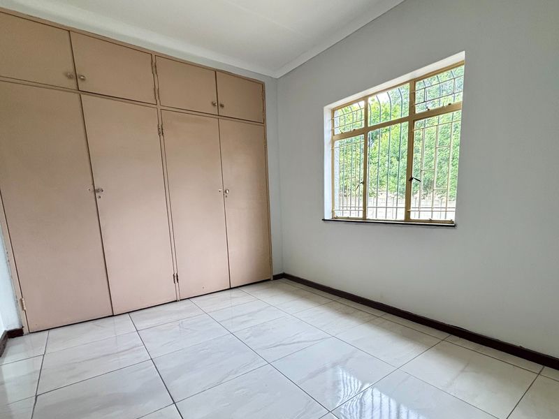 A clean, empty bedroom featuring neutral tiled flooring, built-in cupboards for storage, and a window fitted with security bars.