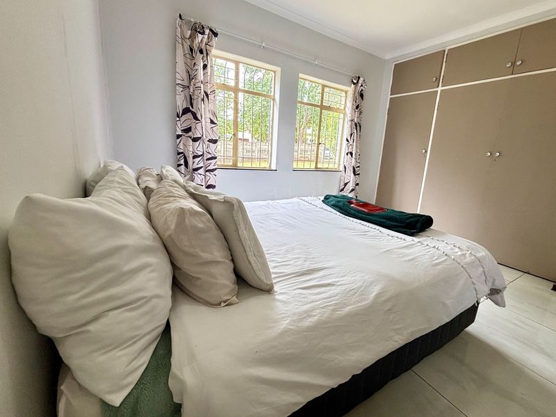 A bedroom featuring built-in cupboards, tiled flooring, and windows fitted with security bars, offering a functional and well-lit living space.