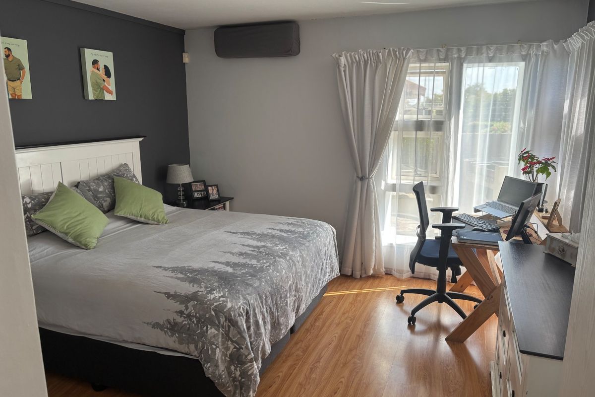 A well-lit bedroom featuring laminate flooring, a wall-mounted air conditioning unit, and large windows with curtains, providing a comfortable and functional living space.