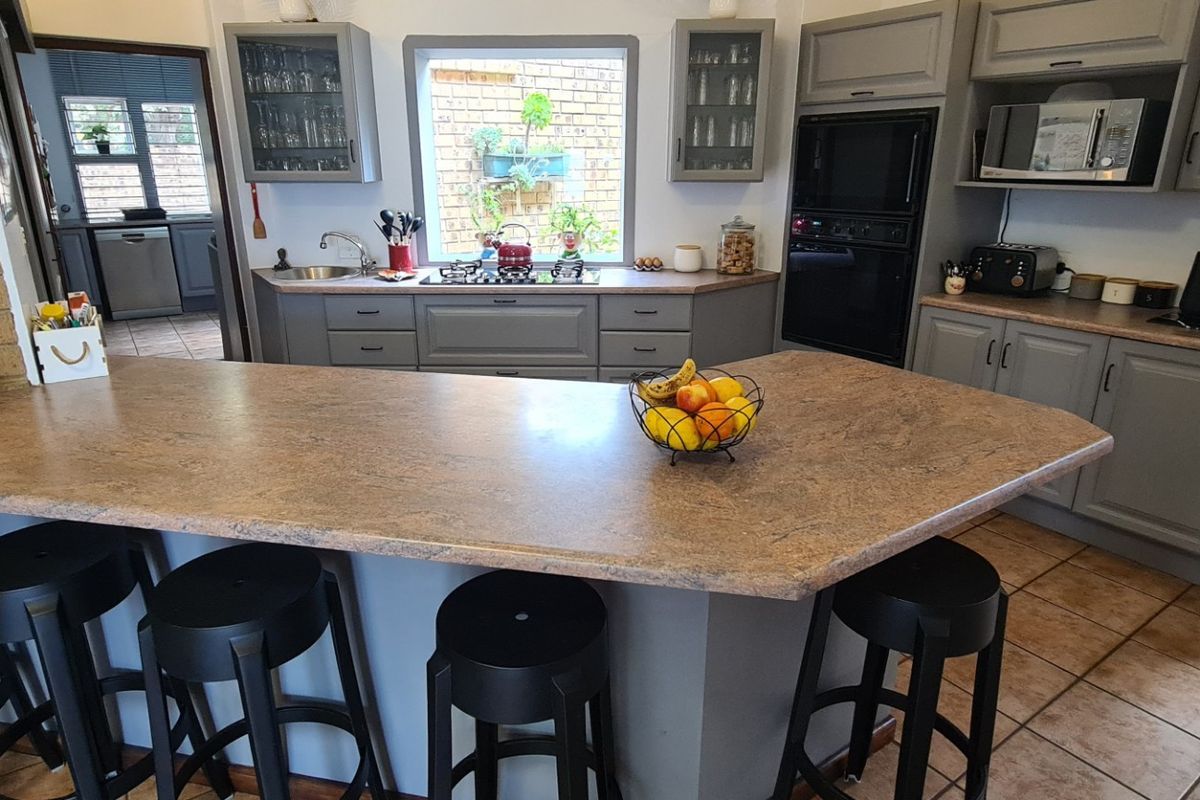 A functional kitchen featuring grey cabinetry, laminate countertops, a breakfast bar, and built-in appliances, complemented by tiled flooring and an exposed brick feature wall.