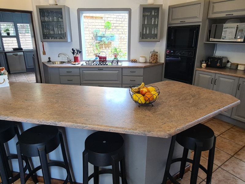 A functional kitchen featuring grey cabinetry, laminate countertops, a breakfast bar, and built-in appliances, complemented by tiled flooring and an exposed brick feature wall.