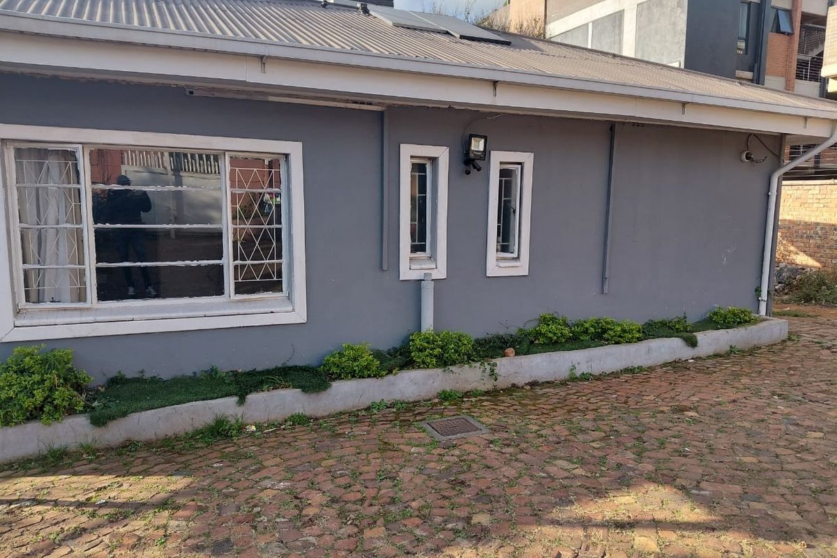 Rear exterior view of the property featuring a corrugated metal roof, security-barred windows, and a cobblestone paved area with a raised garden bed.
