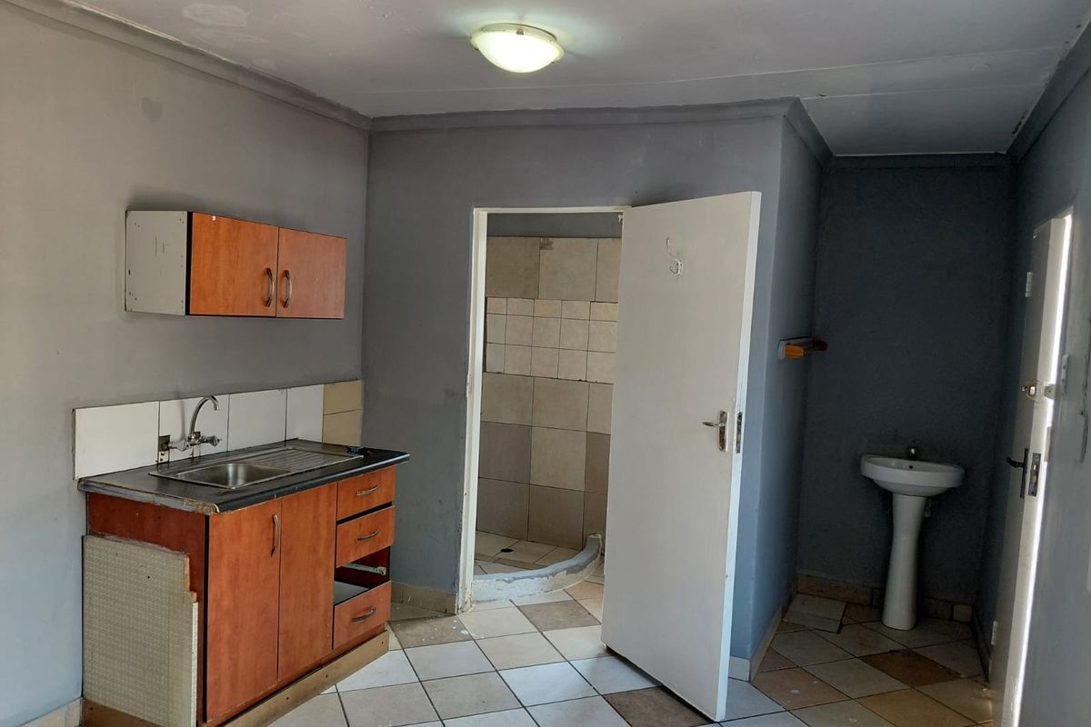 A basic kitchen and bathroom area featuring a small kitchenette, pedestal basin, and tiled shower enclosure, requiring significant cosmetic updates and repairs.