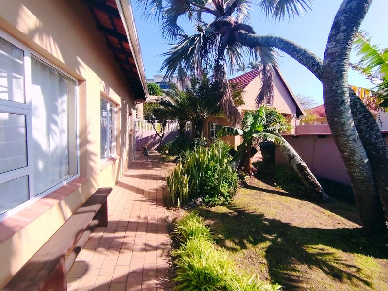 A well-maintained rear exterior area featuring a paved walkway alongside the house, established garden landscaping, and mature palm trees.