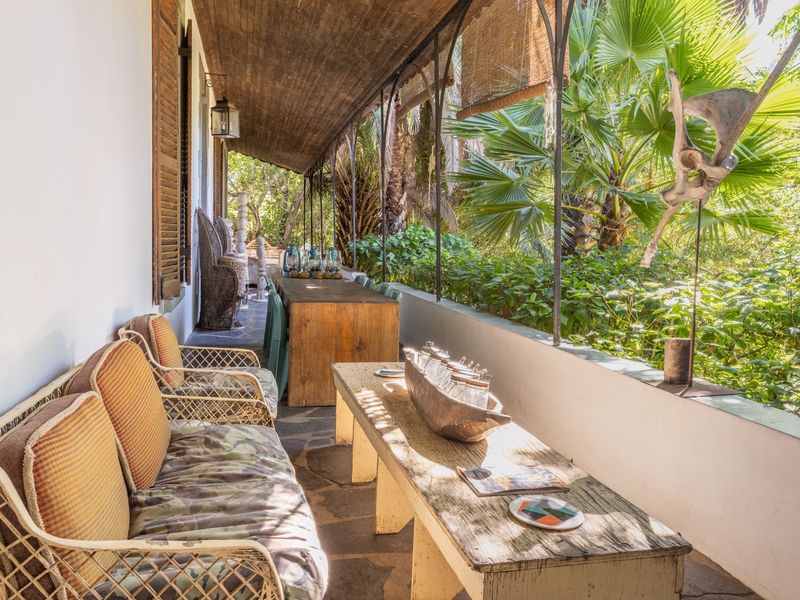 A covered outdoor patio featuring stone flooring, timber ceiling, and wrought iron supports, overlooking a lush garden setting.