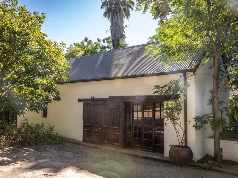 A charming farm-style outbuilding featuring a corrugated metal roof, plastered walls, and rustic sliding timber doors opening onto a garden area.