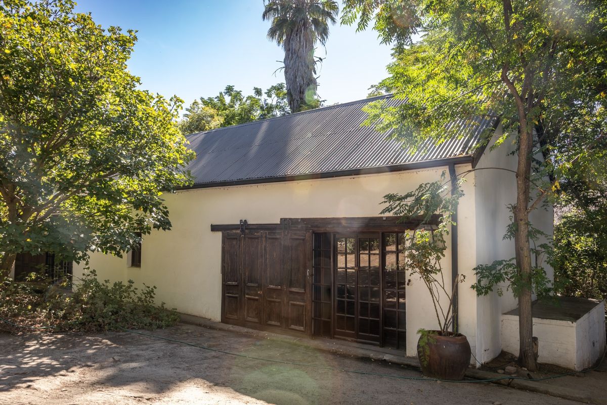 A charming farm-style outbuilding featuring a corrugated metal roof, plastered walls, and rustic sliding timber doors opening onto a garden area.