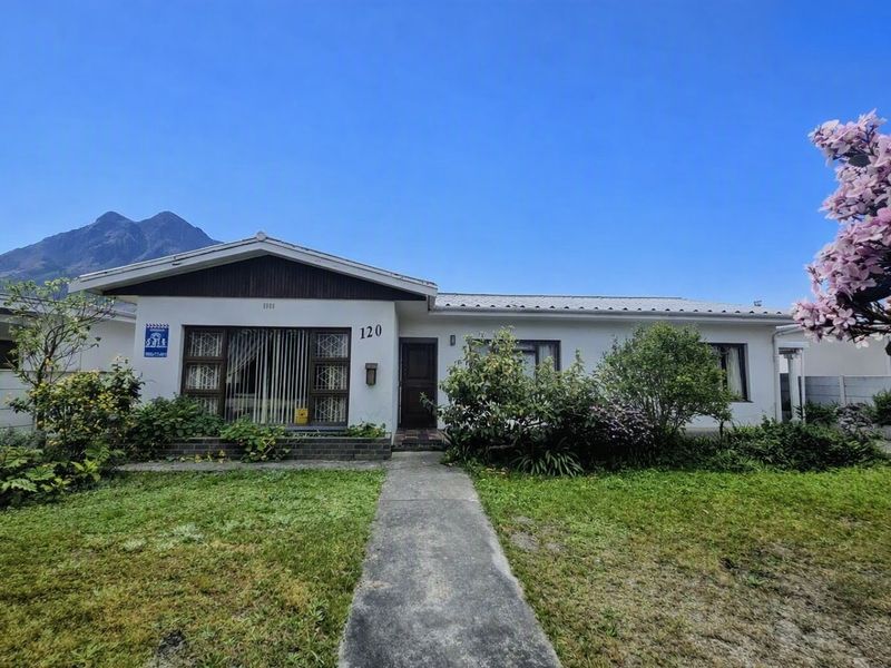 Front elevation of a single-story residential house featuring a concrete walkway, metal roofing, and established garden landscaping against a mountain backdrop.