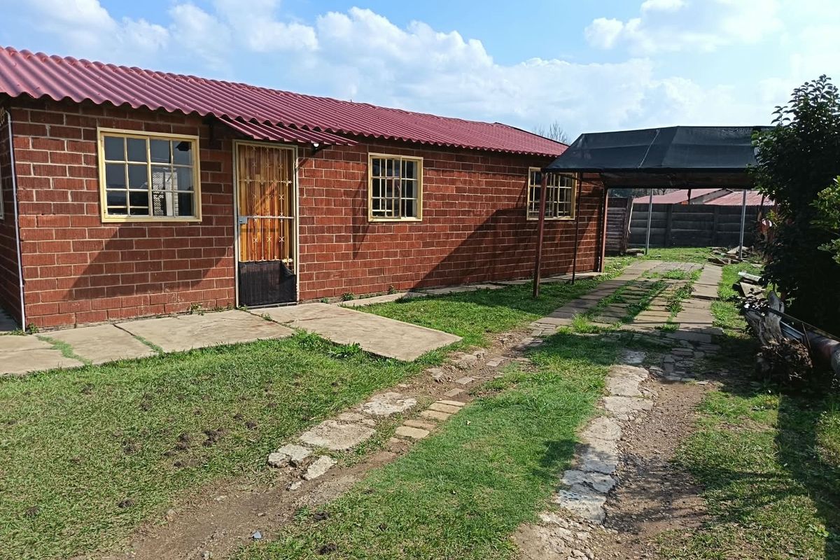 Rear view of a brick house featuring a corrugated metal roof, security gate, and a covered carport area with a paved driveway.