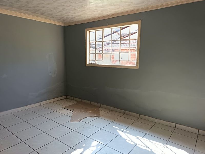 An empty bedroom featuring tiled flooring and a grid-pattern window, currently requiring wall repairs and cosmetic updates to improve its condition.