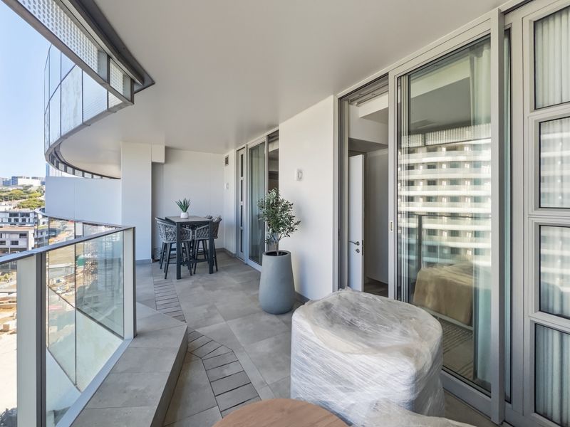 A spacious, modern covered balcony featuring tiled flooring, a sleek glass balustrade, and large sliding glass doors providing seamless indoor-outdoor flow.