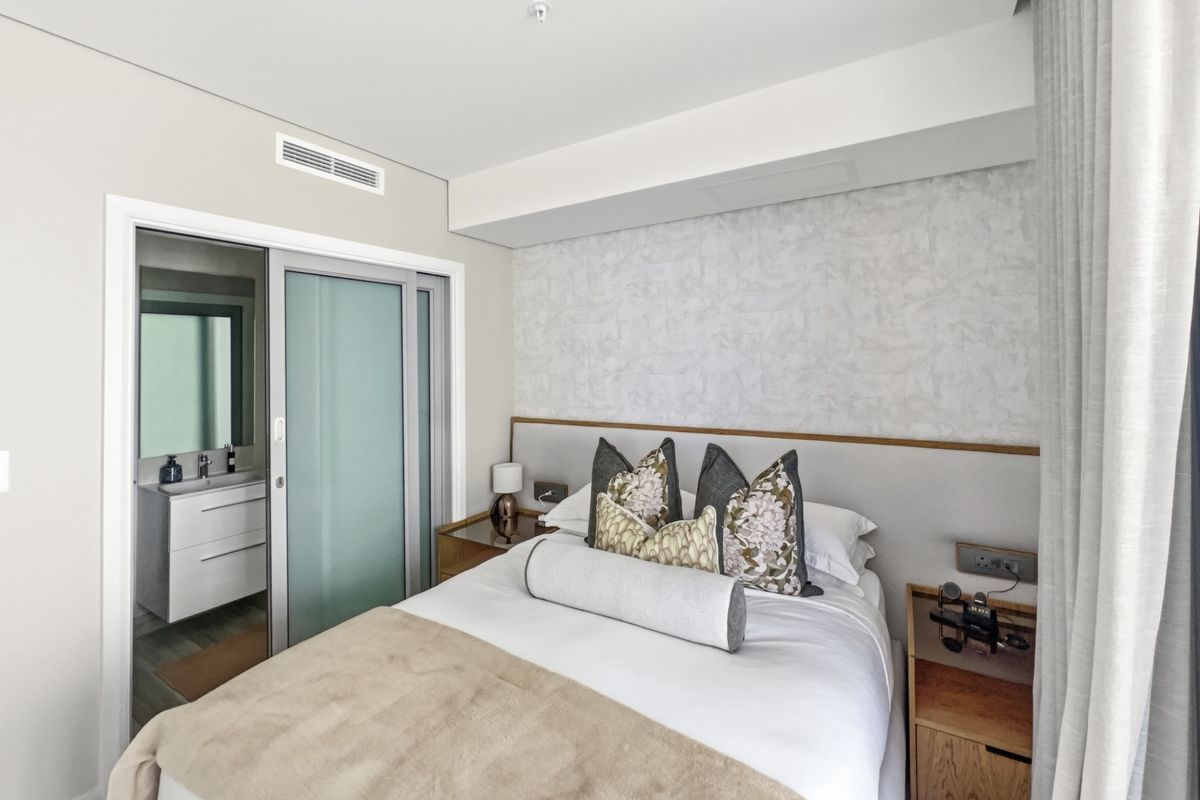 A modern bedroom featuring a built-in headboard, ducted air conditioning, and an adjoining en-suite bathroom with a sliding glass door entry.