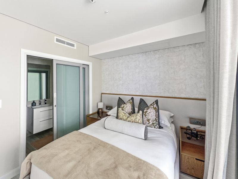 A modern bedroom featuring a built-in headboard, ducted air conditioning, and an adjoining en-suite bathroom with a sliding glass door entry.