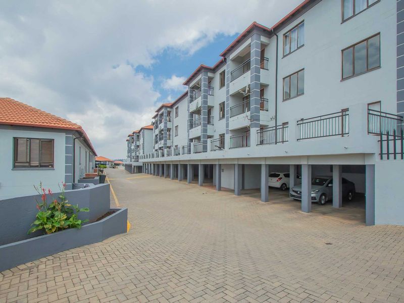 Exterior view of a modern apartment complex featuring covered ground-level parking bays, private balconies, and a paved driveway.