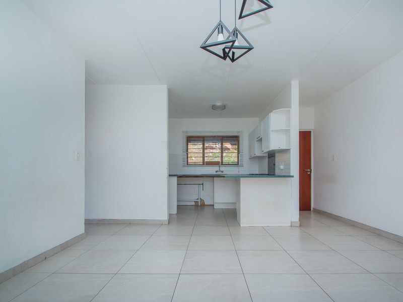 An open-plan living and kitchen area featuring neutral tiled flooring, white cabinetry, and modern geometric pendant lighting in a clean, vacant space.