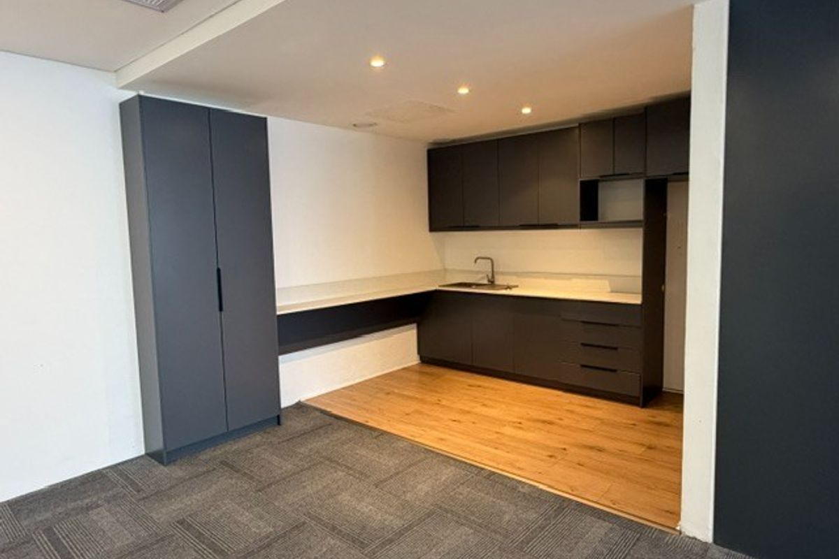 A modern, clean office kitchenette featuring dark grey cabinetry, a stainless steel sink, and a combination of wood-look flooring and carpet tiles.