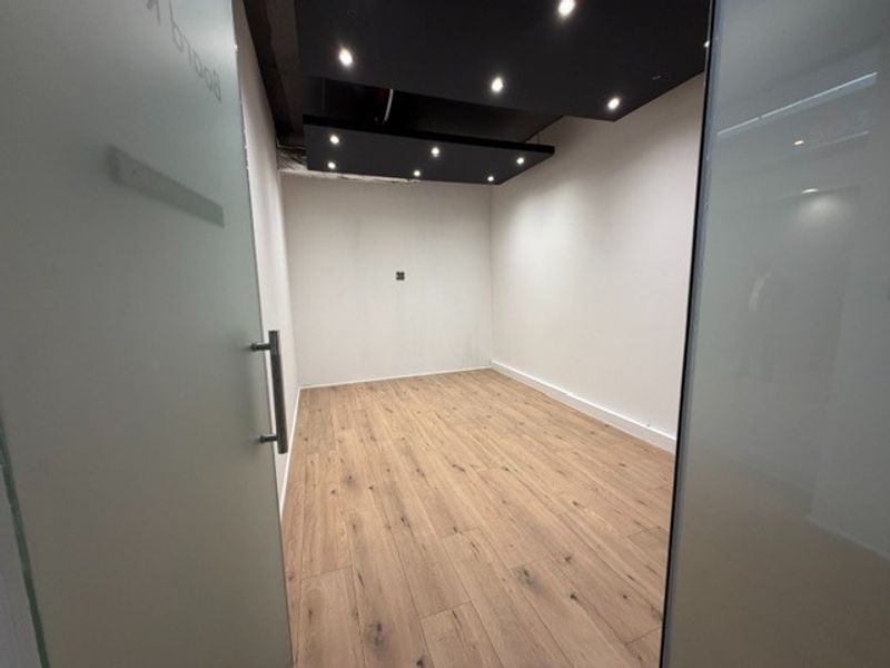 A modern, empty office space featuring wood-look laminate flooring, recessed downlighting, and a sleek frosted glass entrance door.