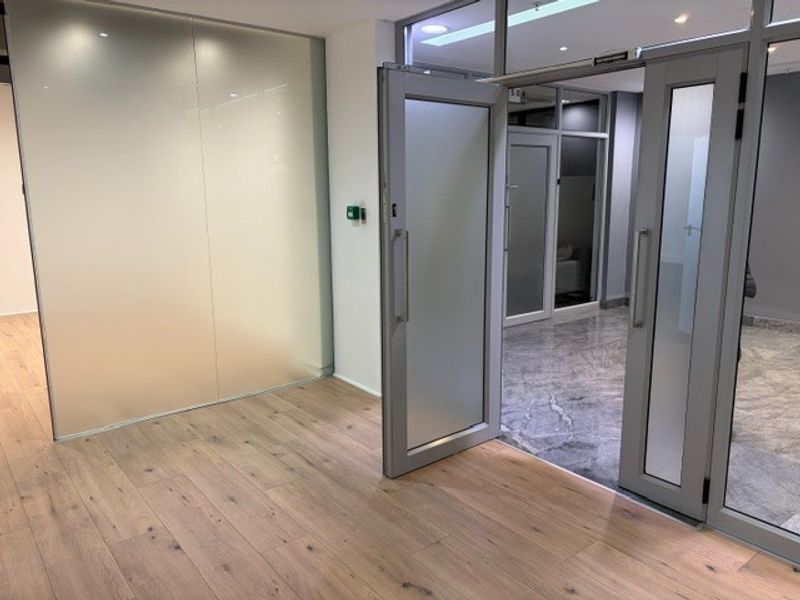 Modern office space featuring high-quality wood laminate flooring, sleek aluminium-framed glass partitions, and professional recessed lighting throughout the interior.